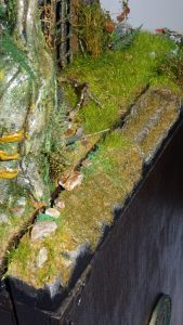 Diorama artesanal detallado de un duende y un pozo estructurado en tres niveles. El nivel superior recrea un jardín exterior exuberante utilizando vegetación natural y preservada (musgos y enredaderas), sobre estancias subterráneas de piedra y madera