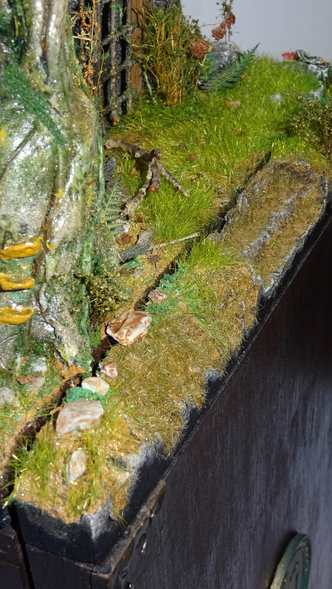 Diorama artesanal detallado de un duende y un pozo estructurado en tres niveles. El nivel superior recrea un jardín exterior exuberante utilizando vegetación natural y preservada (musgos y enredaderas), sobre estancias subterráneas de piedra y madera