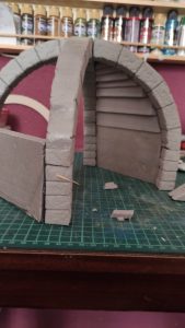 Diorama artesanal detallado de un duende y un pozo estructurado en tres niveles. El nivel superior recrea un jardín exterior exuberante utilizando vegetación natural y preservada (musgos y enredaderas), sobre estancias subterráneas de piedra y madera