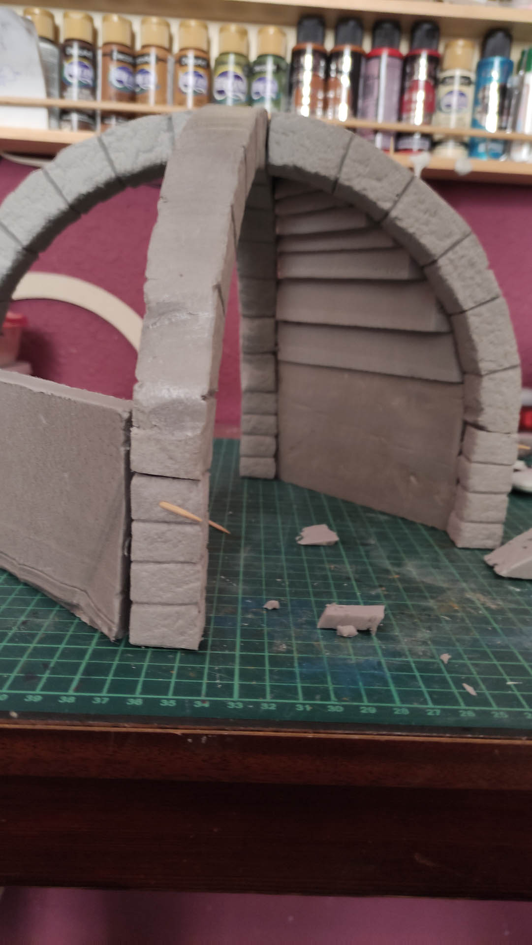 Diorama artesanal detallado de un duende y un pozo estructurado en tres niveles. El nivel superior recrea un jardín exterior exuberante utilizando vegetación natural y preservada (musgos y enredaderas), sobre estancias subterráneas de piedra y madera