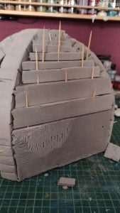 Diorama artesanal detallado de un duende y un pozo estructurado en tres niveles. El nivel superior recrea un jardín exterior exuberante utilizando vegetación natural y preservada (musgos y enredaderas), sobre estancias subterráneas de piedra y madera