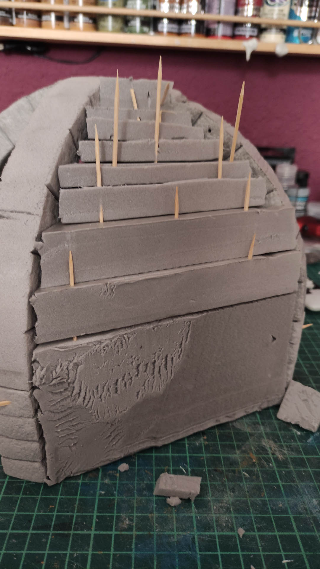 Diorama artesanal detallado de un duende y un pozo estructurado en tres niveles. El nivel superior recrea un jardín exterior exuberante utilizando vegetación natural y preservada (musgos y enredaderas), sobre estancias subterráneas de piedra y madera