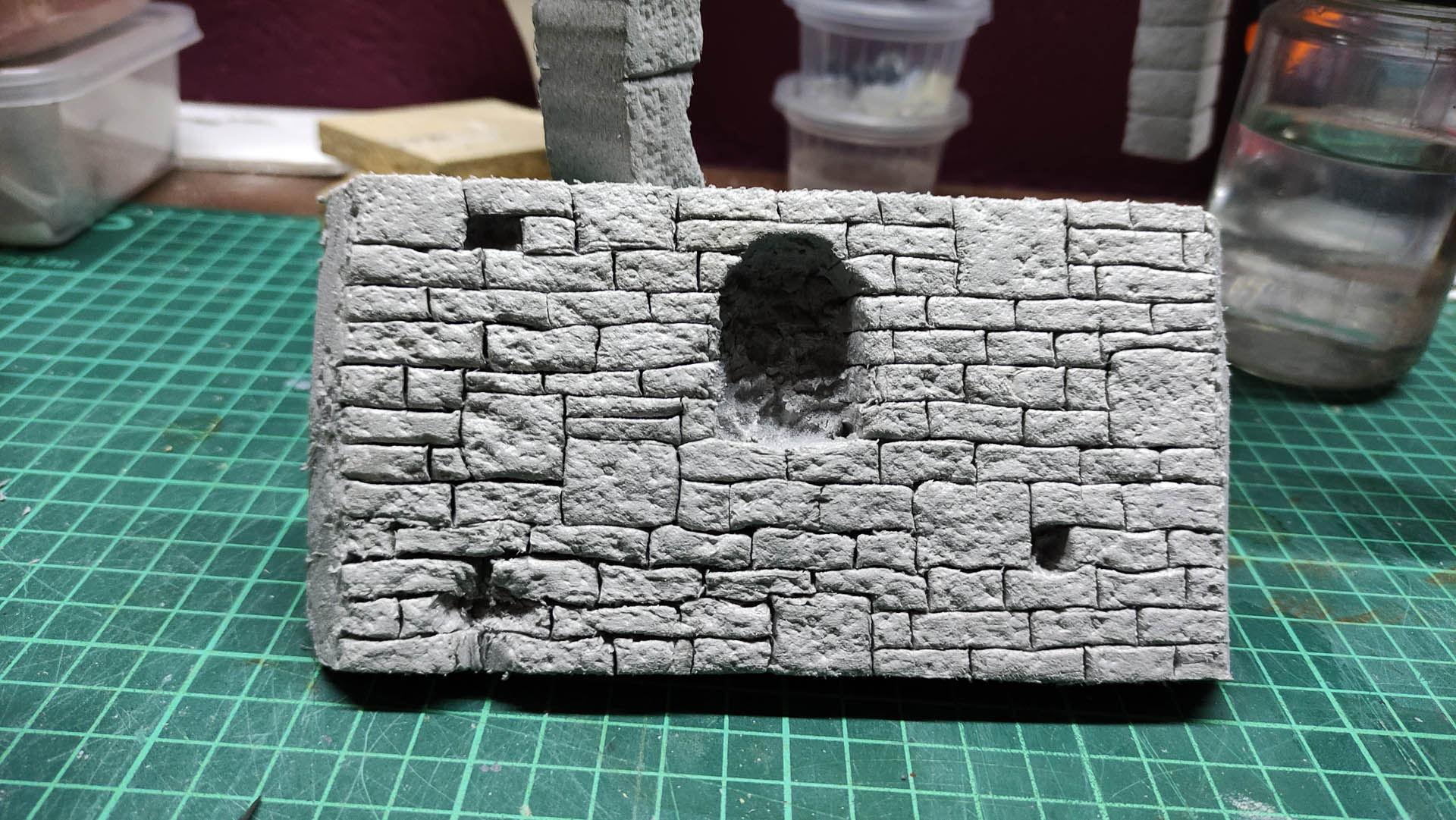 Diorama artesanal detallado de un duende y un pozo estructurado en tres niveles. El nivel superior recrea un jardín exterior exuberante utilizando vegetación natural y preservada (musgos y enredaderas), sobre estancias subterráneas de piedra y madera