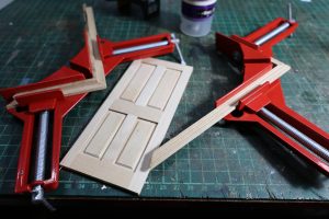 Proceso de construcción del diorama "Las Gemelas": recreación artesanal del pasillo del hotel Overlook con papel pintado geométrico en miniatura, carpintería detallada de puertas de hotel