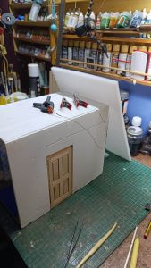 Proceso de construcción del diorama "Las Gemelas": recreación artesanal del pasillo del hotel Overlook con papel pintado geométrico en miniatura, carpintería detallada de puertas de hotel