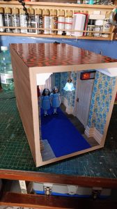 Proceso de construcción del diorama "Las Gemelas": recreación artesanal del pasillo del hotel Overlook con papel pintado geométrico en miniatura, carpintería detallada de puertas de hotel