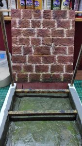 Proceso artesanal del diorama "El Nombre de la Rosa": recreación detallada de los baños del monasterio medieval con Berengario muerto en la bañera, modelado de texturas de piedra húmeda y envejecimiento de mobiliario de madera real