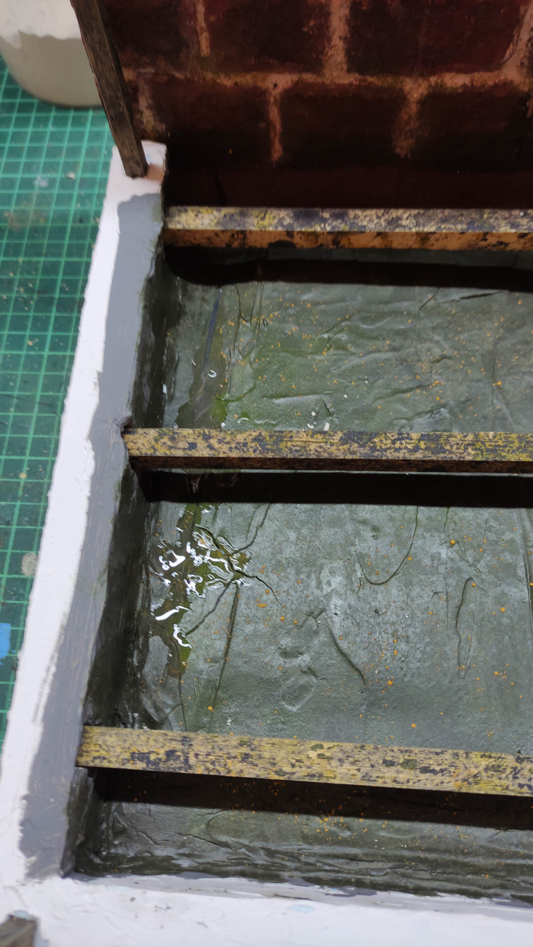 Proceso artesanal del diorama "El Nombre de la Rosa": recreación detallada de los baños del monasterio medieval con Berengario muerto en la bañera, modelado de texturas de piedra húmeda y envejecimiento de mobiliario de madera real