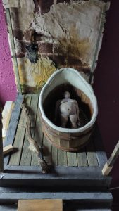 Proceso artesanal del diorama "El Nombre de la Rosa": recreación detallada de los baños del monasterio medieval con Berengario muerto en la bañera, modelado de texturas de piedra húmeda y envejecimiento de mobiliario de madera real