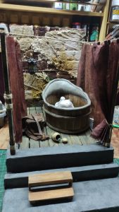 Proceso artesanal del diorama "El Nombre de la Rosa": recreación detallada de los baños del monasterio medieval con Berengario muerto en la bañera, modelado de texturas de piedra húmeda y envejecimiento de mobiliario de madera real