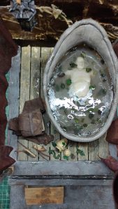 Proceso artesanal del diorama "El Nombre de la Rosa": recreación detallada de los baños del monasterio medieval con Berengario muerto en la bañera, modelado de texturas de piedra húmeda y envejecimiento de mobiliario de madera real