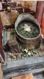Proceso artesanal del diorama "El Nombre de la Rosa": recreación detallada de los baños del monasterio medieval con Berengario muerto en la bañera, modelado de texturas de piedra húmeda y envejecimiento de mobiliario de madera real