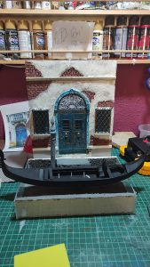 Diorama artesanal de Venecia durante la peste negra, mostrando a dos médicos de la peste con máscara frente a una góndola en un canal, con iluminación de farol.