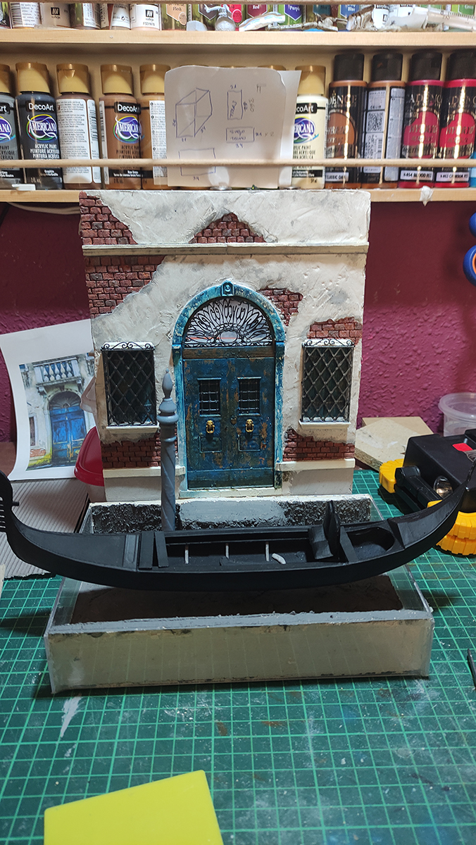 Diorama artesanal de Venecia durante la peste negra, mostrando a dos médicos de la peste con máscara frente a una góndola en un canal, con iluminación de farol.