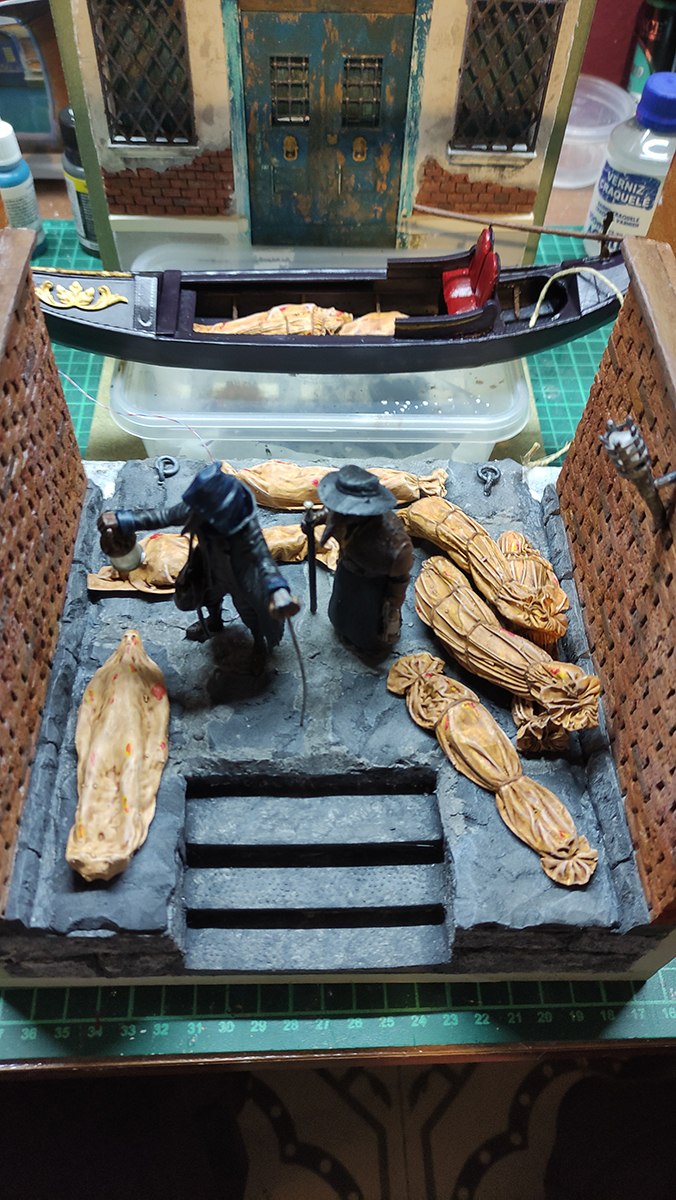 Diorama artesanal de Venecia durante la peste negra, mostrando a dos médicos de la peste con máscara frente a una góndola en un canal, con iluminación de farol.