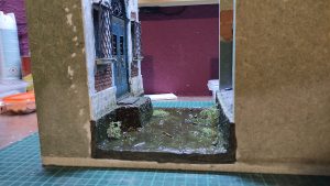 Diorama artesanal de Venecia durante la peste negra, mostrando a dos médicos de la peste con máscara frente a una góndola en un canal, con iluminación de farol.