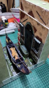 Diorama artesanal de Venecia durante la peste negra, mostrando a dos médicos de la peste con máscara frente a una góndola en un canal, con iluminación de farol.