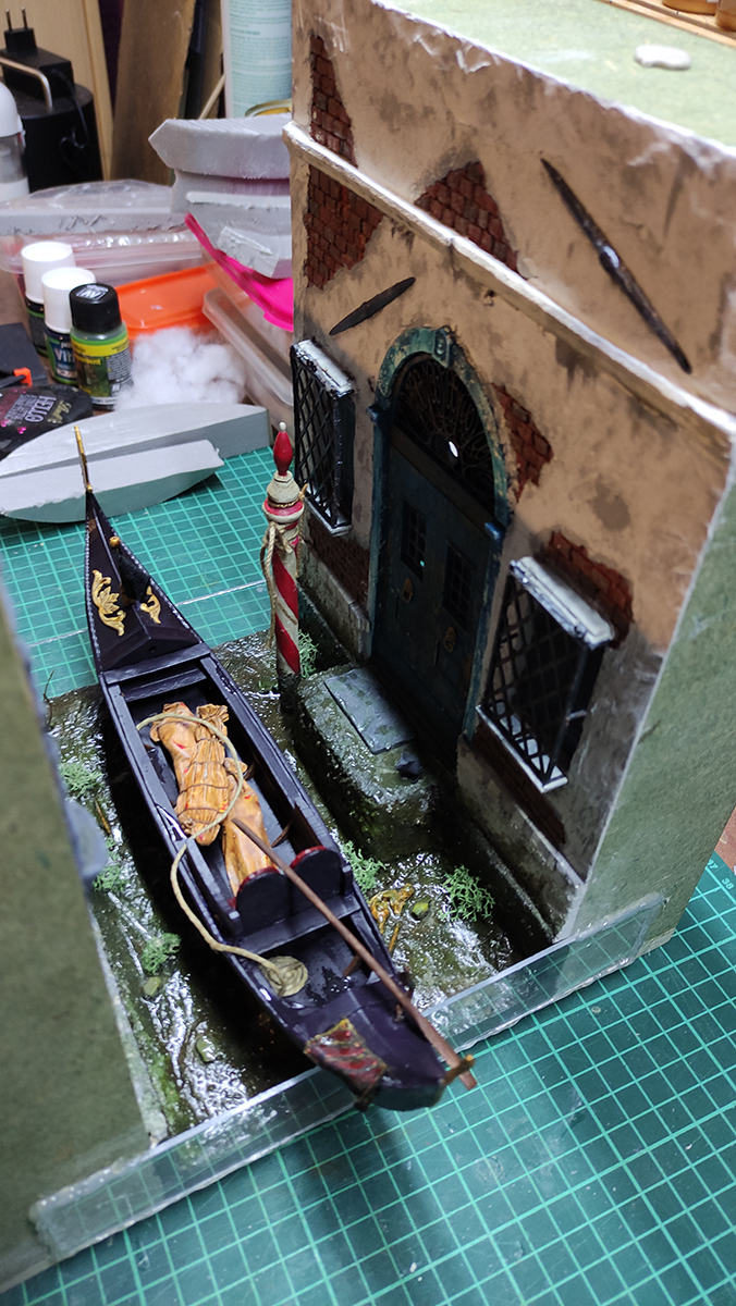 Diorama artesanal de Venecia durante la peste negra, mostrando a dos médicos de la peste con máscara frente a una góndola en un canal, con iluminación de farol.