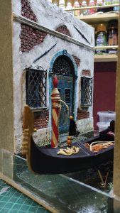 Diorama artesanal de Venecia durante la peste negra, mostrando a dos médicos de la peste con máscara frente a una góndola en un canal, con iluminación de farol.