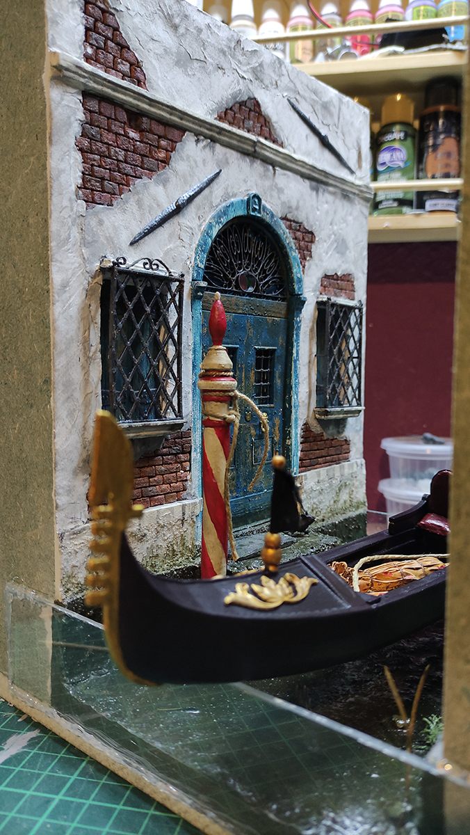 Diorama artesanal de Venecia durante la peste negra, mostrando a dos médicos de la peste con máscara frente a una góndola en un canal, con iluminación de farol.