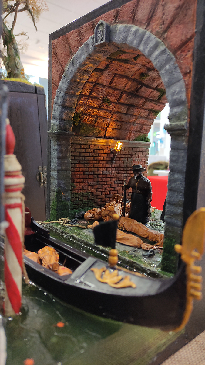 Diorama artesanal de Venecia durante la peste negra, mostrando a dos médicos de la peste con máscara frente a una góndola en un canal, con iluminación de farol.