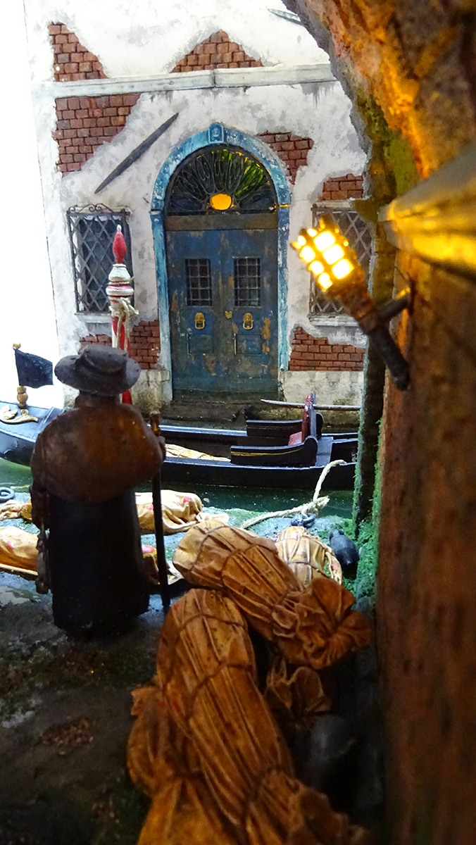 Diorama artesanal de Venecia durante la peste negra, mostrando a dos médicos de la peste con máscara frente a una góndola en un canal, con iluminación de farol.