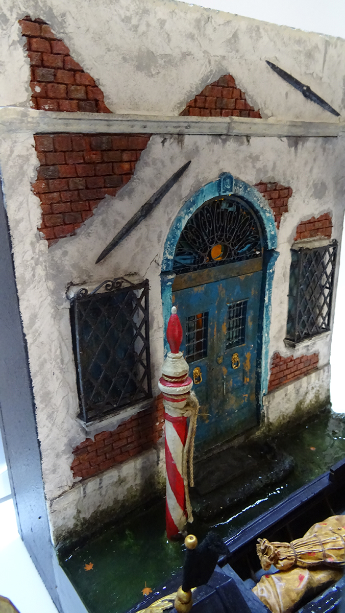 Diorama artesanal de Venecia durante la peste negra, mostrando a dos médicos de la peste con máscara frente a una góndola en un canal, con iluminación de farol.