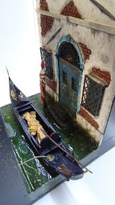 Diorama artesanal de Venecia durante la peste negra, mostrando a dos médicos de la peste con máscara frente a una góndola en un canal, con iluminación de farol.
