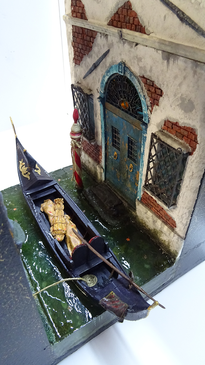 Diorama artesanal de Venecia durante la peste negra, mostrando a dos médicos de la peste con máscara frente a una góndola en un canal, con iluminación de farol.