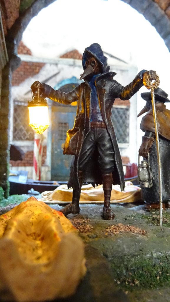 Diorama artesanal de Venecia durante la peste negra, mostrando a dos médicos de la peste con máscara frente a una góndola en un canal, con iluminación de farol.