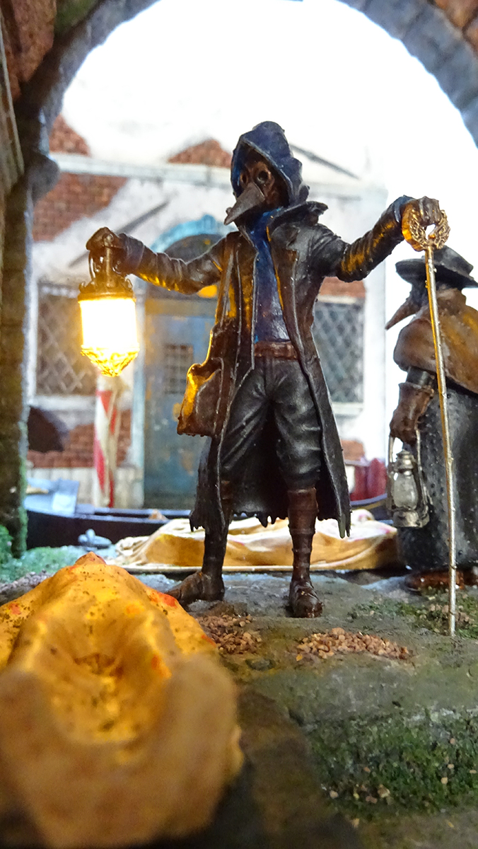 Diorama artesanal de Venecia durante la peste negra, mostrando a dos médicos de la peste con máscara frente a una góndola en un canal, con iluminación de farol.