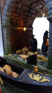 Diorama artesanal de Venecia durante la peste negra, mostrando a dos médicos de la peste con máscara frente a una góndola en un canal, con iluminación de farol.