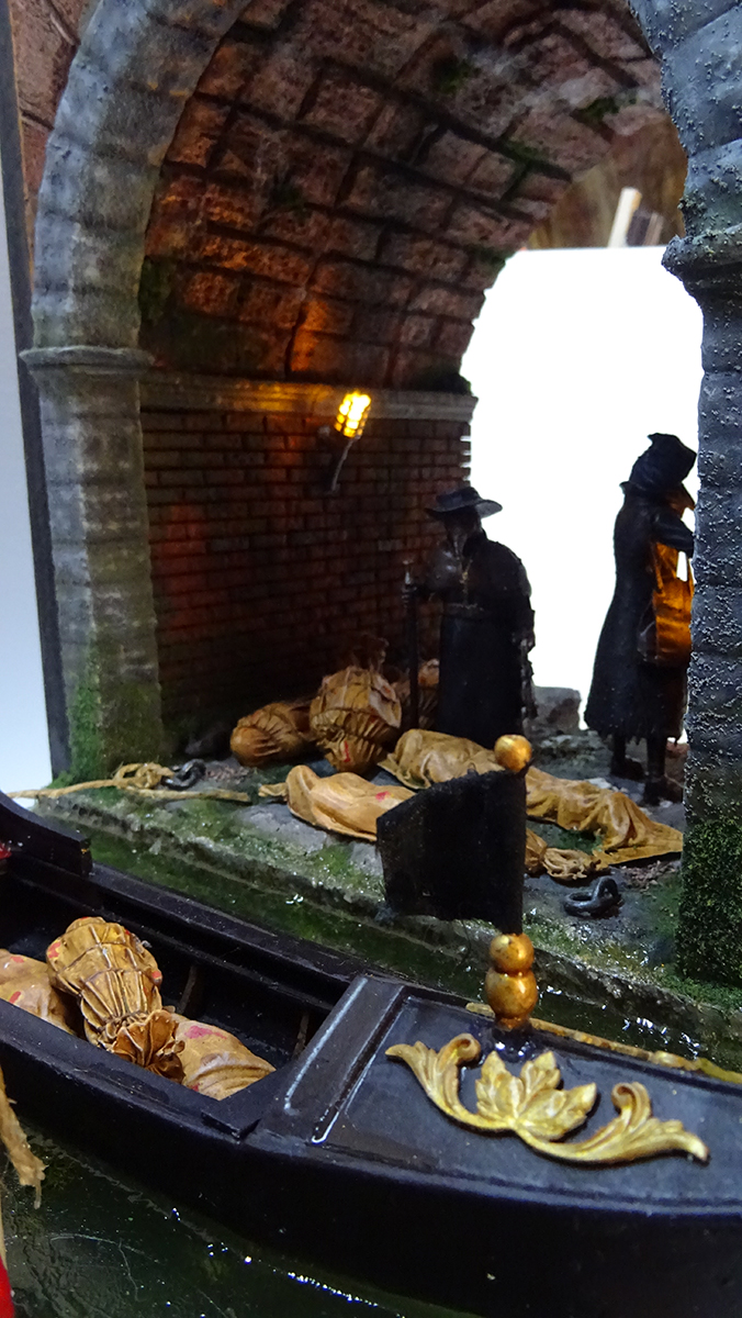 Diorama artesanal de Venecia durante la peste negra, mostrando a dos médicos de la peste con máscara frente a una góndola en un canal, con iluminación de farol.