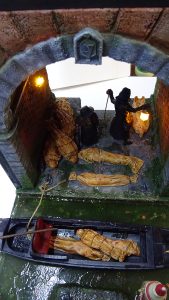 Diorama artesanal de Venecia durante la peste negra, mostrando a dos médicos de la peste con máscara frente a una góndola en un canal, con iluminación de farol.