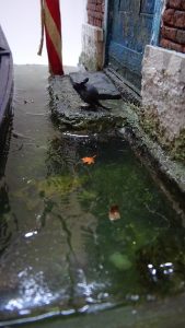 Diorama artesanal de Venecia durante la peste negra, mostrando a dos médicos de la peste con máscara frente a una góndola en un canal, con iluminación de farol.