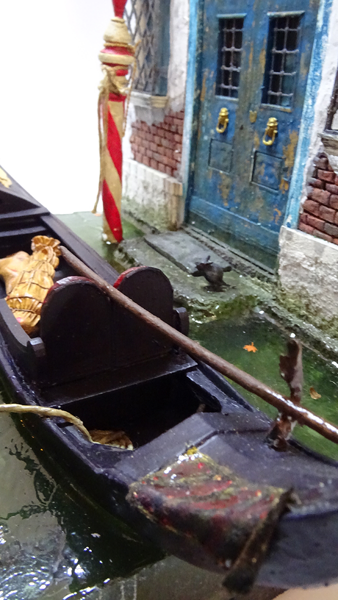 Diorama artesanal de Venecia durante la peste negra, mostrando a dos médicos de la peste con máscara frente a una góndola en un canal, con iluminación de farol.