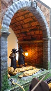 Diorama artesanal de Venecia durante la peste negra, mostrando a dos médicos de la peste con máscara frente a una góndola en un canal, con iluminación de farol.