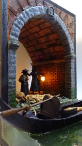 Diorama artesanal de Venecia durante la peste negra, mostrando a dos médicos de la peste con máscara frente a una góndola en un canal, con iluminación de farol.