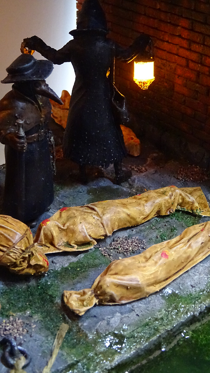 Diorama artesanal de Venecia durante la peste negra, mostrando a dos médicos de la peste con máscara frente a una góndola en un canal, con iluminación de farol.