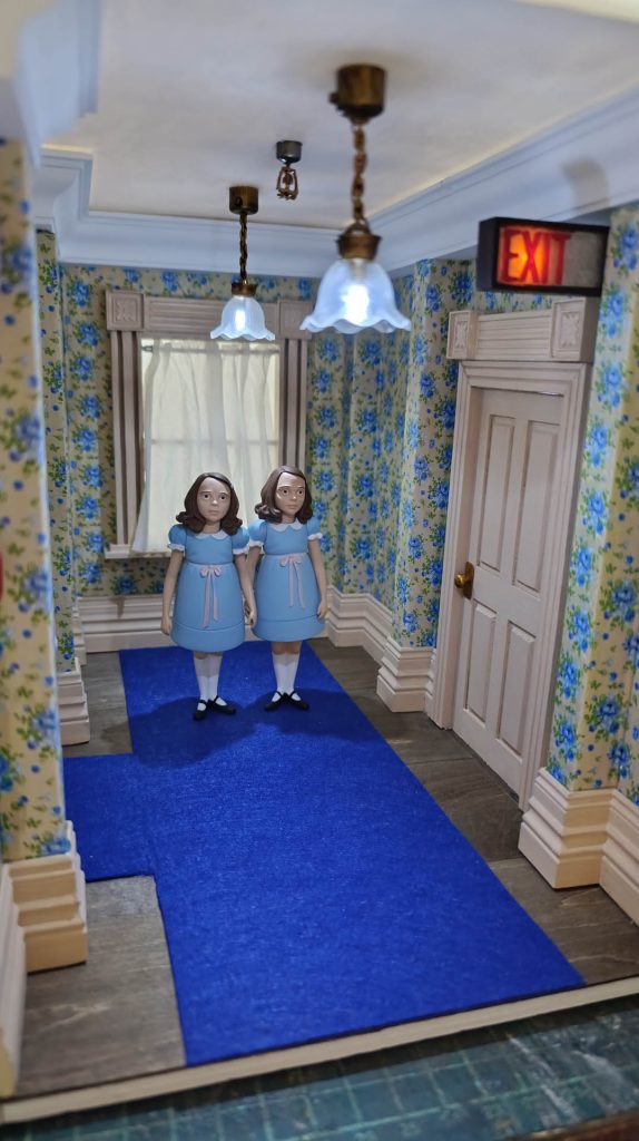 Miniatura artesanal de la escena de las gemelas de El Resplandor. Diorama con iluminación LED real, texturas de papel tapiz victoriano y acabados de alta precisión en madera y resina. Pieza de colección de realismo extremo por 27dioramas.