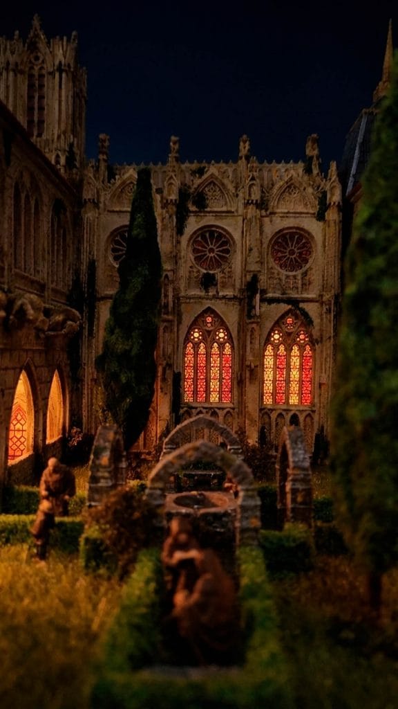 Primer plano en perspectiva del diorama de una abadía gótica, mostrando el claustro iluminado de noche con vidrieras rojas y naranjas.