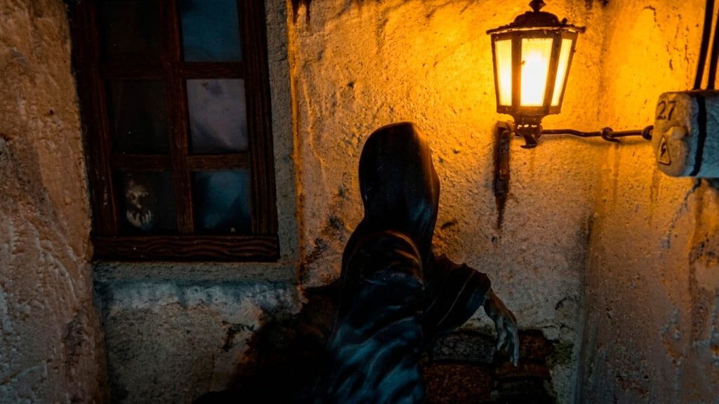 Primer plano de un diorama de terror sobre la leyenda de Saucedilla, mostrando un espectro encapuchado bajo un farol y un rostro terrorífico tras la ventana.