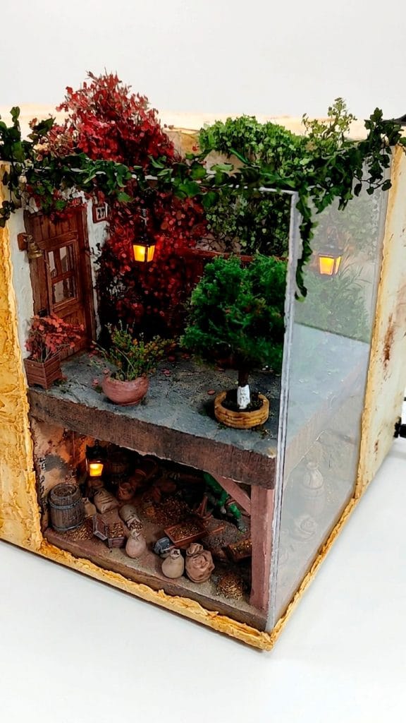 Diorama artesanal detallado de un duende bajo un patio con vegetación densa y una estancia subterránea con barriles y sacos, iluminado con faroles LED cálidos.