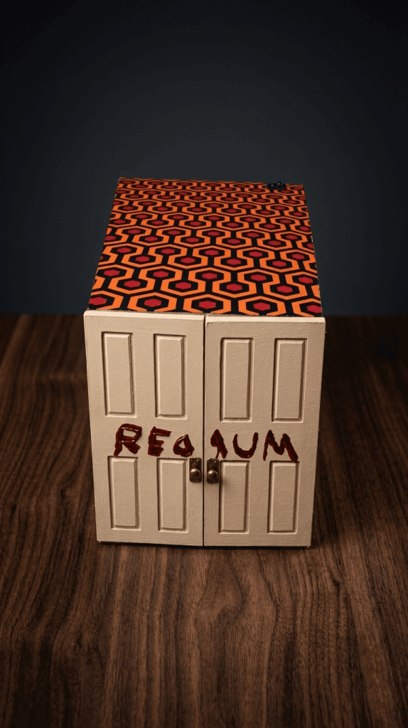 Diorama de El Resplandor con puertas cerradas y mensaje REDRUM sobre caja con estampado de alfombra del Hotel Overlook.