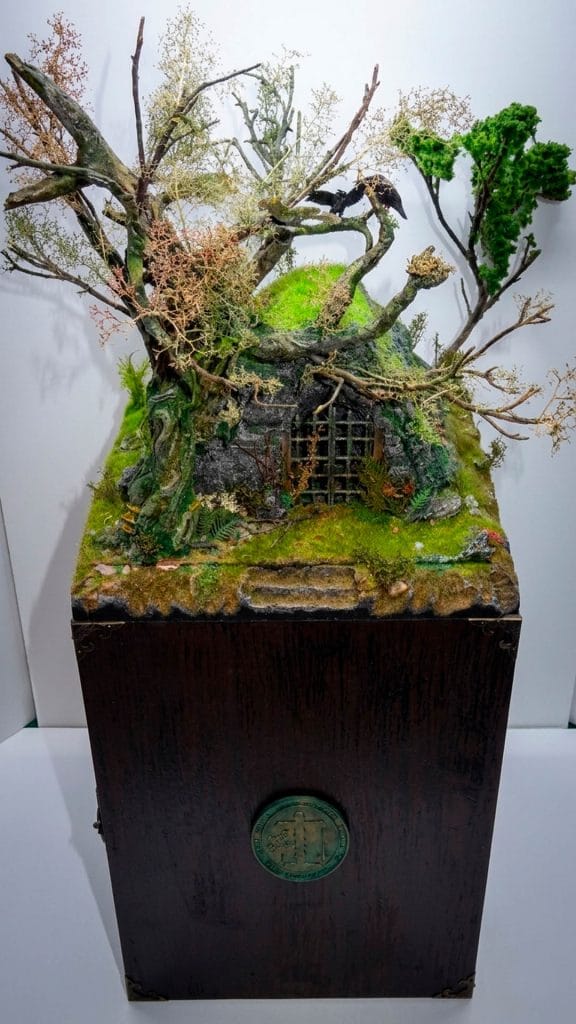 Diorama artesanal de la Inquisición que muestra una entrada a una mazmorra oculta bajo un túmulo con vegetación, árboles secos y cuervos.