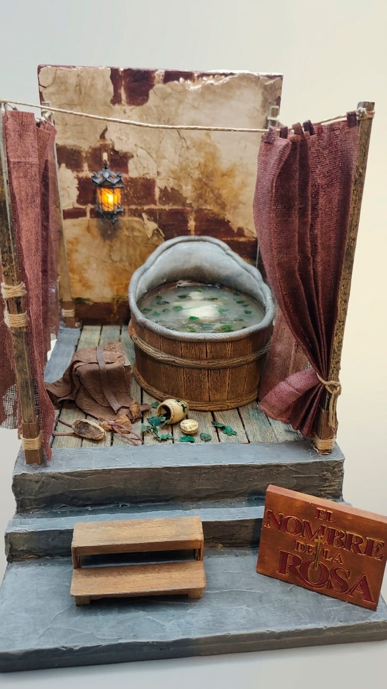 Diorama artesanal de 'El Nombre de la Rosa' que recrea la muerte de Berengario en la tina de los baños, con cortinas de tela, iluminación de farol y placa identificativa.