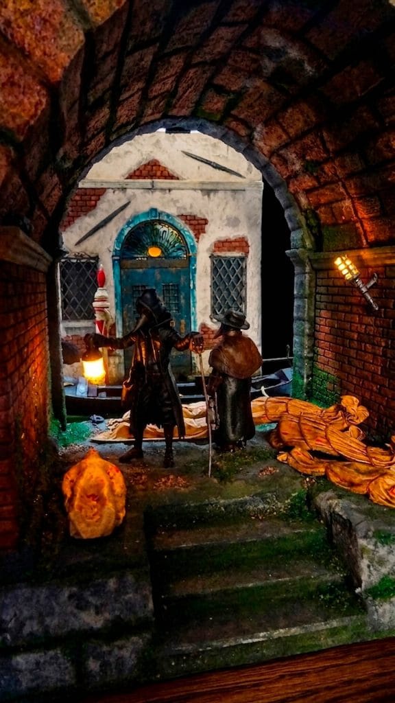 Diorama artesanal de Venecia durante la peste negra, mostrando a dos médicos de la peste con máscara frente a una góndola en un canal, con iluminación de farol.