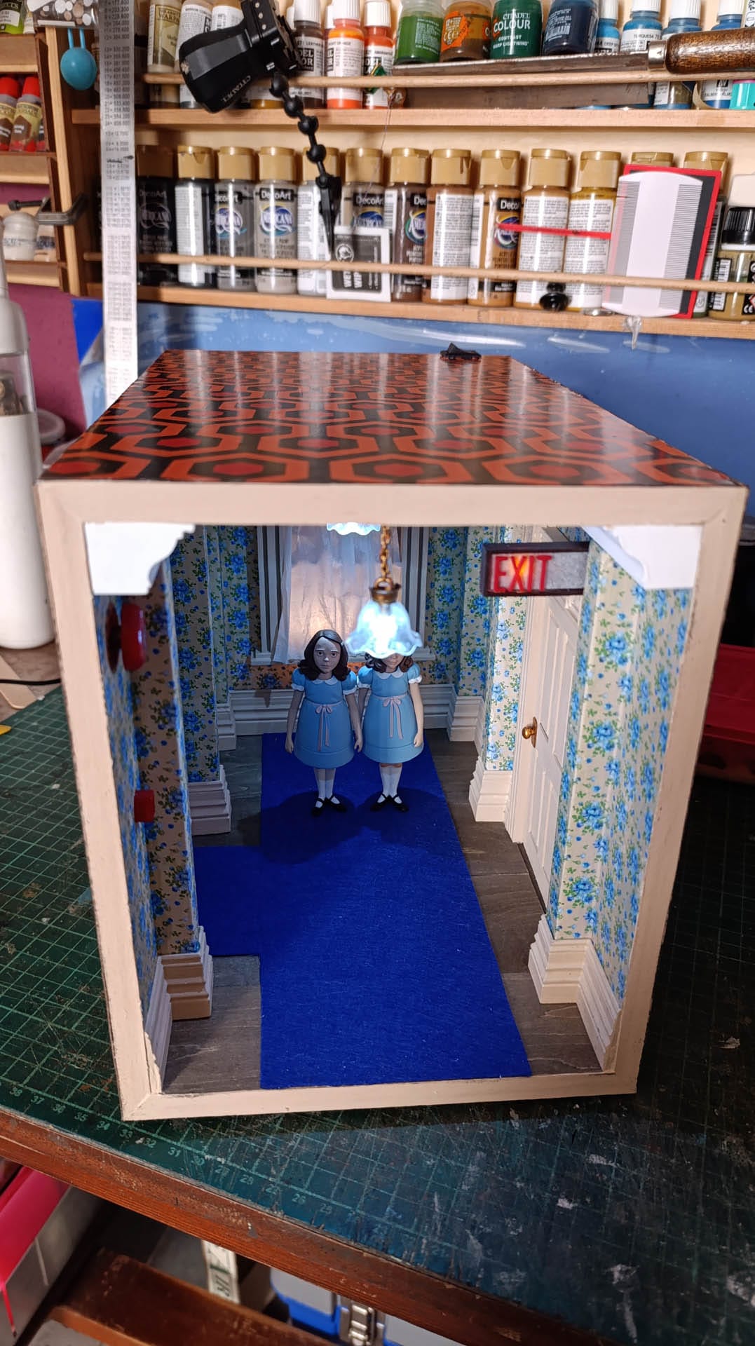 Proceso de construcción del diorama "Las Gemelas": recreación artesanal del pasillo del hotel Overlook con papel pintado geométrico en miniatura, carpintería detallada de puertas de hotel