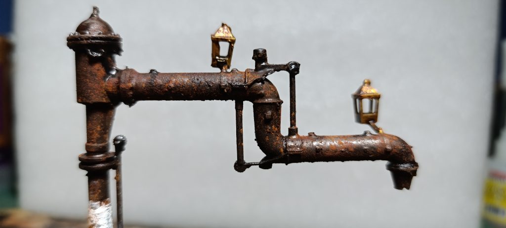 Primer plano de una grúa de agua ferroviaria en miniatura con un intenso acabado de óxido corroído. Dos pequeñas linternas metálicas de latón cuelgan de la tubería horizontal superior. Una sección de pintura blanca es visible en la columna vertical. El fondo está desenfocado.