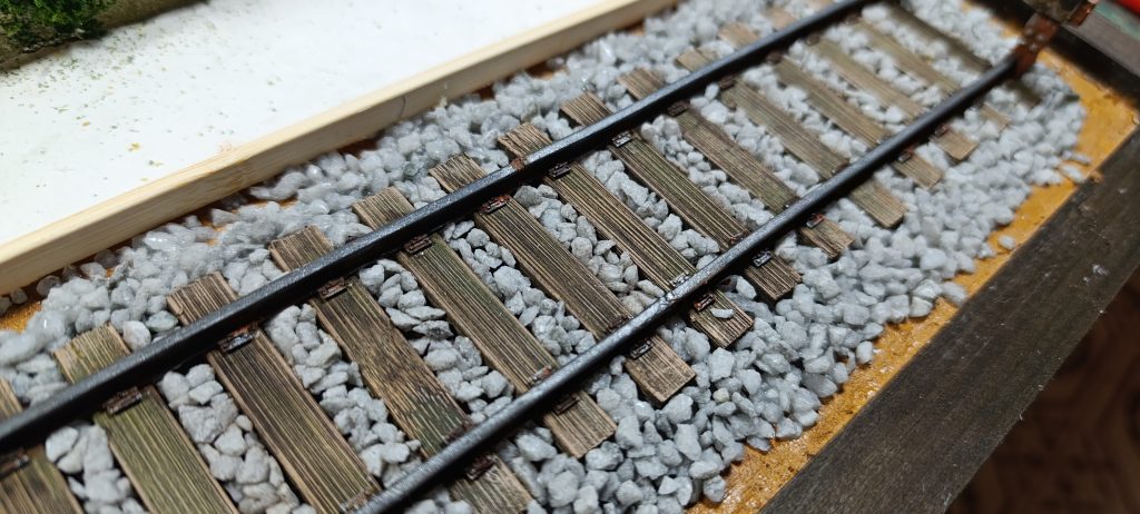 Primer plano cenital de vías ferroviarias en miniatura con traviesas de madera oscura y envejecida. Los raíles metálicos están pintados de negro. La vía descansa sobre un lecho de piedras reales de color gris claro que forma el balasto. En la esquina superior izquierda se ve una sección de terreno sin terminar.